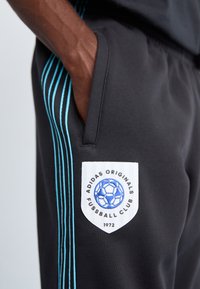 Sorte treningbukser med blå vertikale striper, med et skjoldformet brodert logo med en fotball og tekst. Myk stoff.