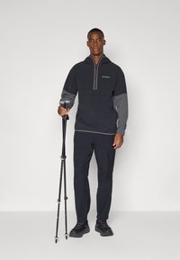 Schwarzer Quarter-Zip-Pullover mit grauen Akzenten, kombiniert mit schwarzen Hosen. Das Model hält Trekkingstöcke und zeigt somit einen Outdoor-Style.