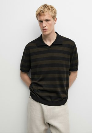 Jeune homme aux cheveux blonds courts portant une chemise à manches courtes rayée noire et vert foncé et un pantalon beige clair, debout contre un mur blanc.