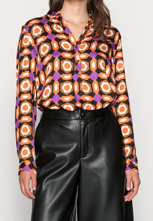 Femme portant un chemisier à manches longues avec des motifs géométriques orange, violet et blanc, rentré dans un pantalon en cuir noir taille haute.