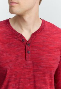 Rød bomulds henley skjorte med et struktureret mønster, en to-knaps placket og en ribbet krave. Subtile variationer øger den visuelle interesse.