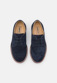 Chaussures en daim bleu marine à lacets avec un motif brogue, doublure en cuir beige et couture blanche contrastante le long de la semelle.