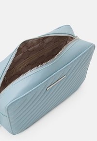 Borsa a tracolla trapuntata blu chiaro con un esterno liscio, chiusura con zip argentata e un pannello con logo in rilievo. Gli interni presentano una foderatura marrone.