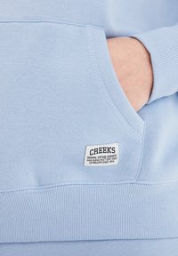 Hellblaues Sweatshirt mit geripptem Saum und Bündchen, ausgestattet mit einer Fronttasche und einem gewebten Etikett mit "CREEKS" und Textdetails.