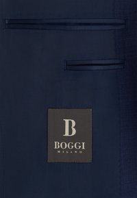 Tissu bleu foncé avec deux poches à rabat horizontales et une étiquette noire cousue portant l'inscription "B BOGGI MILANO" en texte blanc.