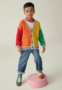 Mehrfarbige gestrickte Strickjacke mit orangefarbenen, roten, gelben und grünen Segmenten, ausgestattet mit einem V-Ausschnitt, gestreiften Bündchen und schwarzen Knöpfen. Blaue Jeans und bunte Sneakers.