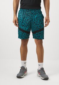 Shorts pour hommes en bleu sarcelle avec un motif léopard noir, dotés d'accents en tissu noir et d'une taille élastique. Associés à des chaussures de sport grises.