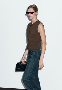 Haut en tricot sans manches de couleur marron avec une poche à l'avant, associé à un jean en denim foncé, tenant un petit sac à main tissé noir. Design minimaliste.