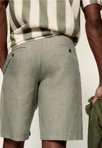 Uomo che indossa pantaloni corti in lino verde chiaro con tasche posteriori con bottone e una camicia beige e verde a righe infilata dentro, tiene in mano un tessuto verde.