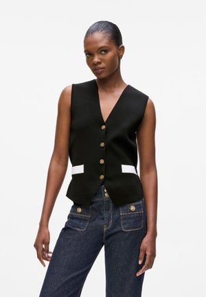 Black sleeveless vest with a deep V-neck, gold buttons, and white accents on pockets. Worn with dark denim jeans featuring gold hardware.