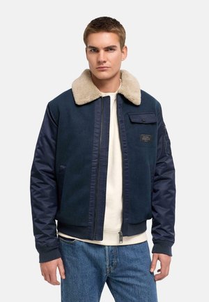 Jeune homme portant une veste bomber bleu marine avec un col en shearling beige, chemise blanche et jean bleu, debout devant un fond uni.