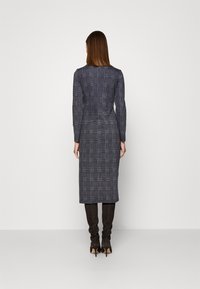 Robe à manches longues de couleur gris foncé avec un large motif à carreaux, design ajusté, longueur genou et coupe droite. Portée avec des bottes à talons hauts.