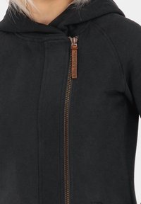 Sudadera con cremallera negra hecha de tela suave, que presenta una capucha ajustable, una cremallera de tono dorado y una pequeña etiqueta de cuero en el tirador de la cremallera.