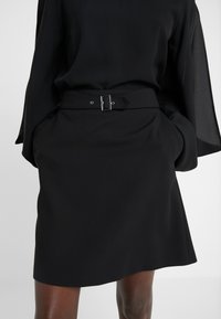 Musta hame, jossa on istuva vyötärö, ja siinä on vyö suorakaiteen muotoisella soljella. Sujuva kangas, polvimittainen, sivutaskuilla ja liehuva muoto.