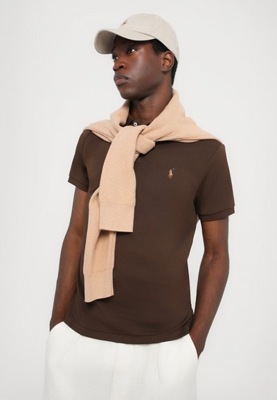 Camisa polo marrón de manga corta con un logo sutil. Suéter beige enrollado alrededor de los hombros, gorra clara a juego. Pantalones blancos.
