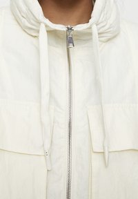 Lekka kurtka z kapturem w kolorze ecru. Posiada przedni zamek błyskawiczny, sznurek ściągający oraz dwie duże kieszenie na piersi. Materiał o fakturze z matowym wykończeniem.