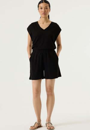 Zwart V-hals top met kapmouwen, gecombineerd met gestructureerde zwarte shorts. De outfit heeft een losse pasvorm en zijzakken. Platte sandalen.