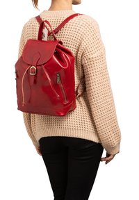 Sac à dos en cuir rouge à la forme arrondie, avec des poches latérales zippées et une fermeture à rabat. Porté par une personne, associé à un pull en laine beige.