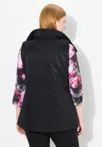 Schwarze, puffy Weste mit hohem Kragen, aus glänzendem Material. Die Rückseite ist glatt, während die Ärmel ein mehrfarbiges Blumenmuster aufweisen.
