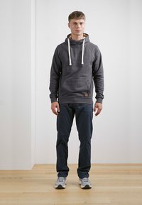 Mørkegrå hættejakke med hvide snore, forlommes og logo-patch; sat sammen med marineblå bukser og grå sneakers. Ensfarvet baggrund.