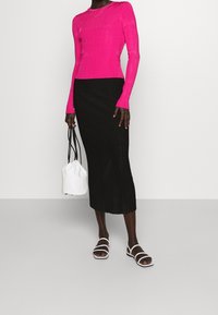 Maglietta aderente a maniche lunghe di un brillante colore rosa con lavorazione a costine abbinata a una gonna midi nera plissettata. Una borsa a secchiello bianca e sandali bianchi con cinturini completano l'outfit.