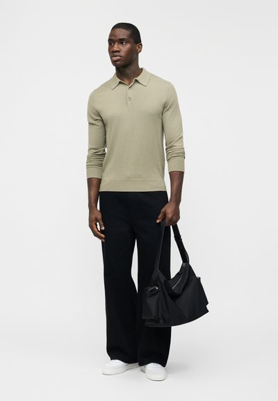 Homme portant un polo beige à manches longues, pantalon noir à jambes larges, baskets blanches, tenant un sac à bandoulière noir, debout devant un fond clair uni.