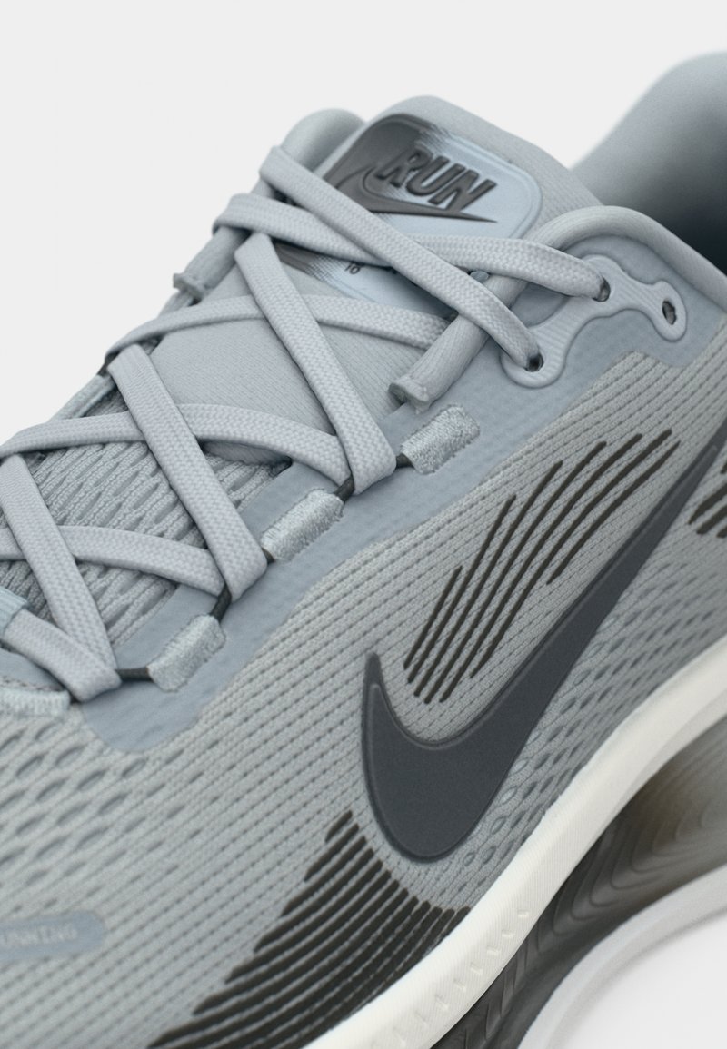 Graue Nike-Laufschuhe mit einem strukturierten Mesh-Obermaterial, schwarzem Logo und flachen Schnürsenkeln. Die Sohle verfügt über eine weiße Zwischensohle und ein gemustertes Traktionsdesign.