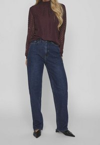 Blusa bordeaux a maniche lunghe con collo alto e motivo a zigzag, abbinata a jeans in denim blu scuro a vita alta. Scarpe nere a punta.