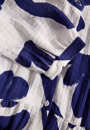 Weißer Stoff mit großen blauen abstrakten Mustern, ausgestattet mit einem geknöpften Kragen und strukturierter Oberfläche. Beinhaltet eine Knopfdetaillierung und Falten.