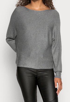 Vrouw draagt een grijze geribde gebreide trui met lange mouwen en zwarte leren broek, staand met één hand achter haar rug.