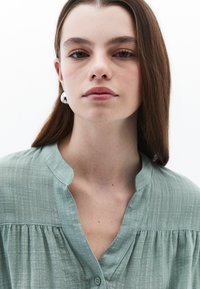 Blusa verde chiaro, dal morbido tessuto, con colletto alla cinese e dettagli plissettati; chiusure a bottone visibili sullo scollo.