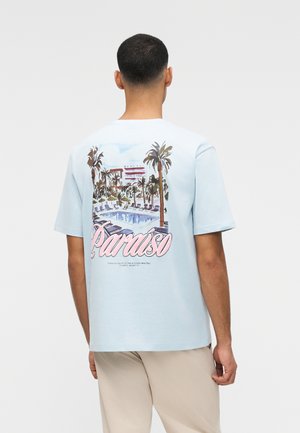 Mann trägt ein hellblaues T-Shirt mit einer bunten Poolszene und dem Wort "Paraiso" auf der Rückseite, kombiniert mit beigen Hosen.