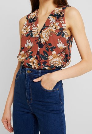 Femme portant un top sans manches à motifs floraux dans des tons rouille et beige, associé à un jean taille haute bleu foncé, main dans la poche.