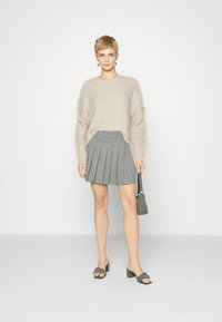 Hellbeige, flauschiger Pullover mit langen Ärmeln, kombiniert mit einem grauen Faltenrock. Das Model hält eine kleine graue Handtasche und trägt graue Absatzschuhe.