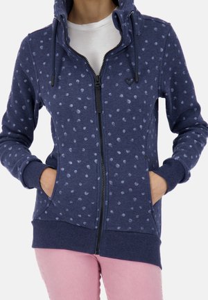 Vrouw draagt een marineblauwe hoodie met ritssluiting, lichtblauwe stippen, zakken en een klein hartdetail, gecombineerd met lichtroze broek.