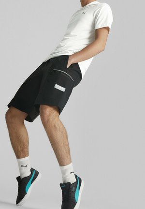 Homme portant un t-shirt blanc Puma, un short cargo noir avec une poche zippée, des chaussettes blanches Puma et des baskets noires Puma avec des rayures bleues sur un fond uni.