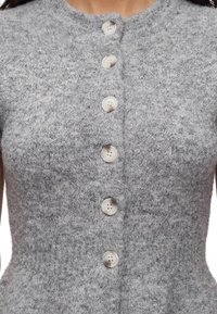 Gros plan d'un cardigan gris tricoté avec six grands boutons blancs au centre, porté par une personne.