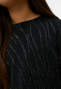 Juodas megztinis su sidabro spalvos metalinėmis siūlėmis banguotu raštu. Audinys turi tekstūruotą išvaizdą ir klasikinę apvalią iškirptę.