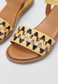 Sandale en cuir jaune avec une large bride à motifs présentant des formes triangulaires noires, bronze et argent ; semelle en caoutchouc ; texture lisse.