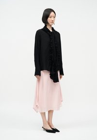Mujer con cabello corto y negro, vistiendo una blusa negra con volantes, falda midi rosa pálido y zapatos de tacón negros con correa en el talón, posando frente a un fondo blanco.