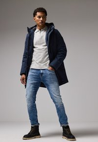 Chaqueta acolchada azul marino, camiseta polo gris, jeans azul claro y botas negras con suelas beige; el modelo está de pie con los brazos relajados, fondo neutro.