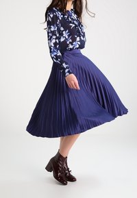 Plisseret, knælængde, mørkeblå nederdel med glat tekstur, kombineret med en langærmet blomsterbluse og blanke brune ankelstøvler.