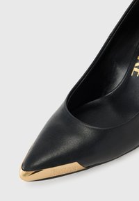 Juodos odinės aukštakulnės bateliai su smailiu nosiu ir auksiniu metaliniu galiuku, pasižymintys tekstūrinėmis apdailomis ir graviruotu logotipu šone.