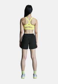 Geel sportbeha met verstelbare bandjes en een uitgesneden rug, gecombineerd met zwarte shorts en grijze hardloopschoenen met neonaccenten.