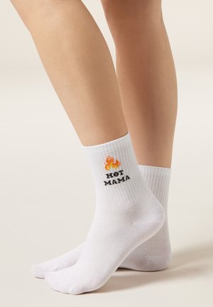 Chaussettes blanches côtelées aux pieds avec un graphisme de flammes et le texte "HOT MAMA" au-dessus de la cheville sur un fond uni.