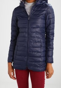Mujer vistiendo una chaqueta acolchada ajustada de color azul marino con cremallera y capucha, y leggings rojos, de pie contra un fondo liso.