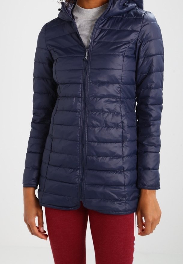 Mujer vistiendo una chaqueta acolchada ajustada de color azul marino con cremallera y capucha, y leggings rojos, de pie contra un fondo liso.