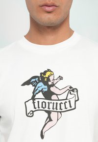 Biela bavlnená tričko s farebnou grafikou anjela s modrými krídlami a čiernym oblečením, doplnené čiernym kurzívnym bannerom "florrucci".