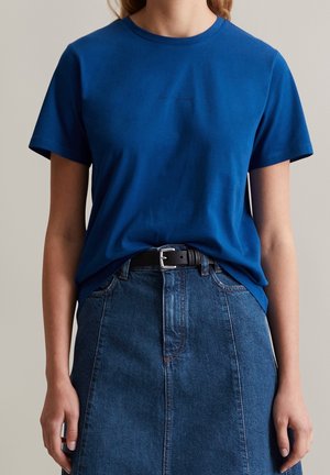 Frau trägt ein schlichtes blaues T-Shirt, das in einen hoch taillierten Jeansrock mit einem schwarzen Gürtel gesteckt ist, und steht vor einem einfarbigen Hintergrund.