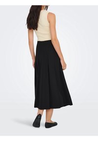 Femme portant un haut beige sans manches, une jupe midi plissée noire et des chaussures plates noires, debout de dos sur un fond uni.